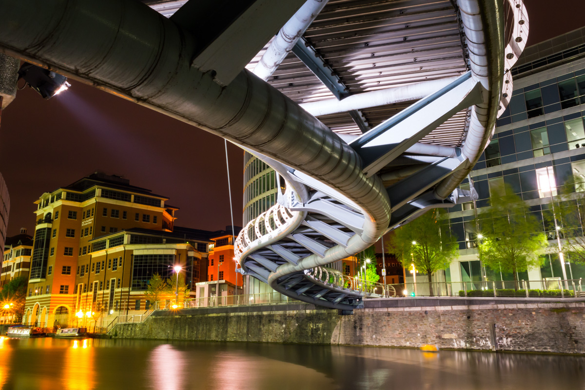 An artistically framed photograph of the underside of the Temple Quay Bridge in the city of Bristol. The photograph is taken at night, with the bridge and buildings in the background illuminated by spot lights.